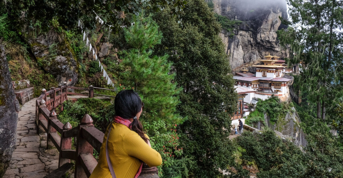 Wide shot, Taktsang, Bhutan. Woman on mountain bridge watches Taktsang. Day.
